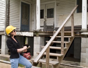 A woman in a hard hat, holding a clipboard, standing in front of an old house.